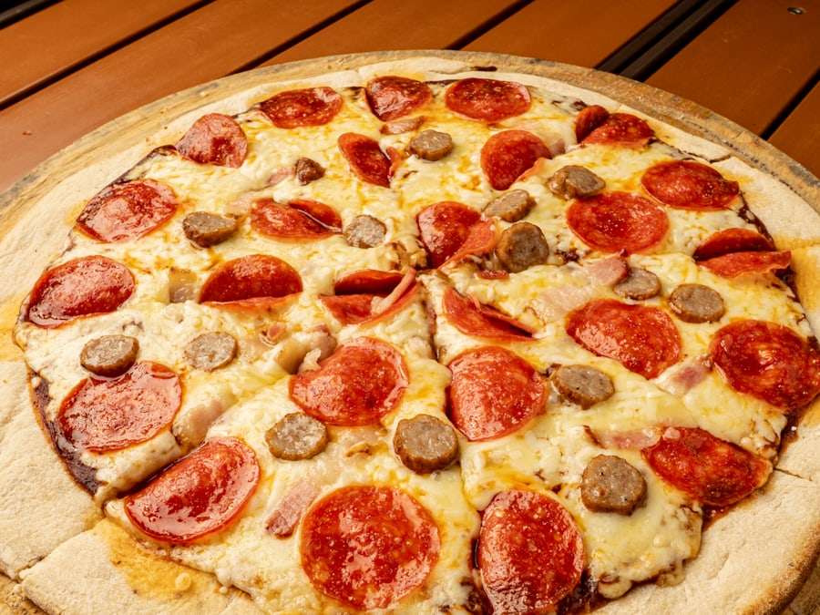 Pizza Meat Lovers de Pizzalicious con pepperoni, salchicha y tocino sobre tabla de madera, vista desde arriba