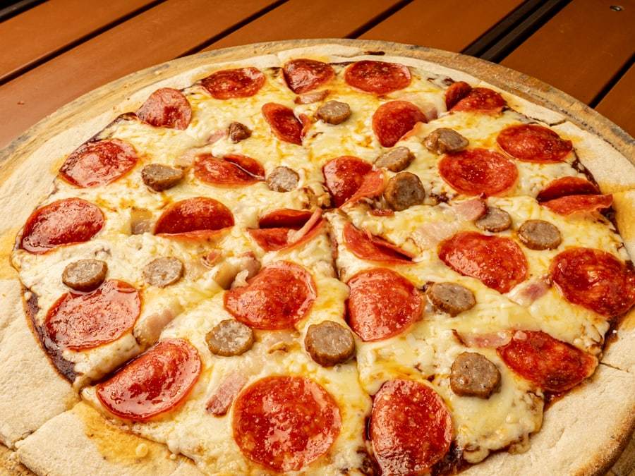 Pizza Meat Lovers de Pizzalicious con pepperoni, salchicha y tocino sobre tabla de madera, vista desde arriba